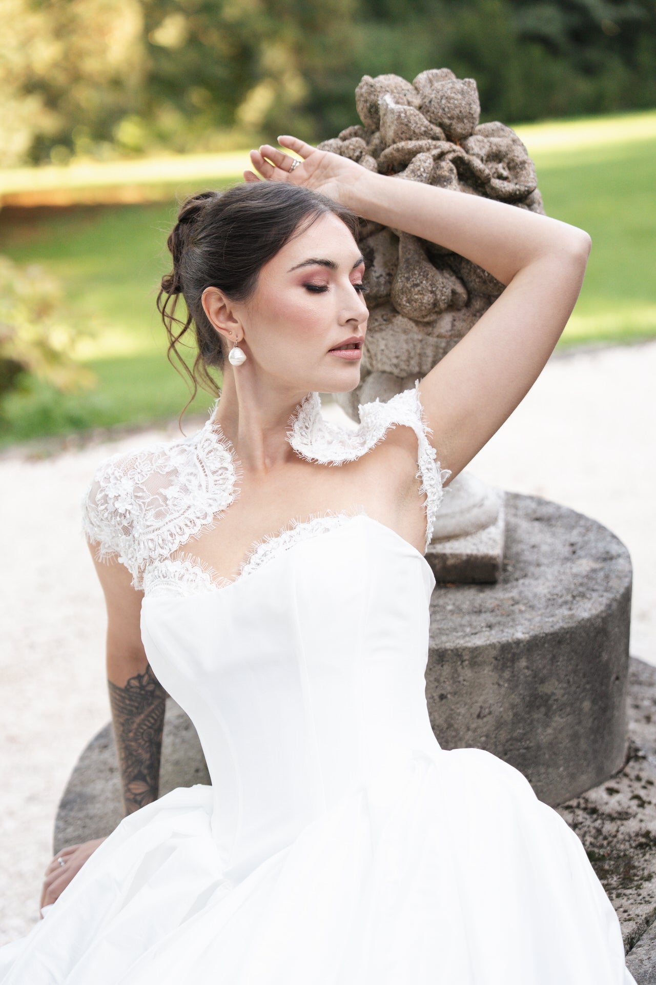 Braut in weißem Kleid mit Spitzenärmeln, sitzt vor steinerner Skulptur im Garten, eleganter Stil, entspannte Pose.