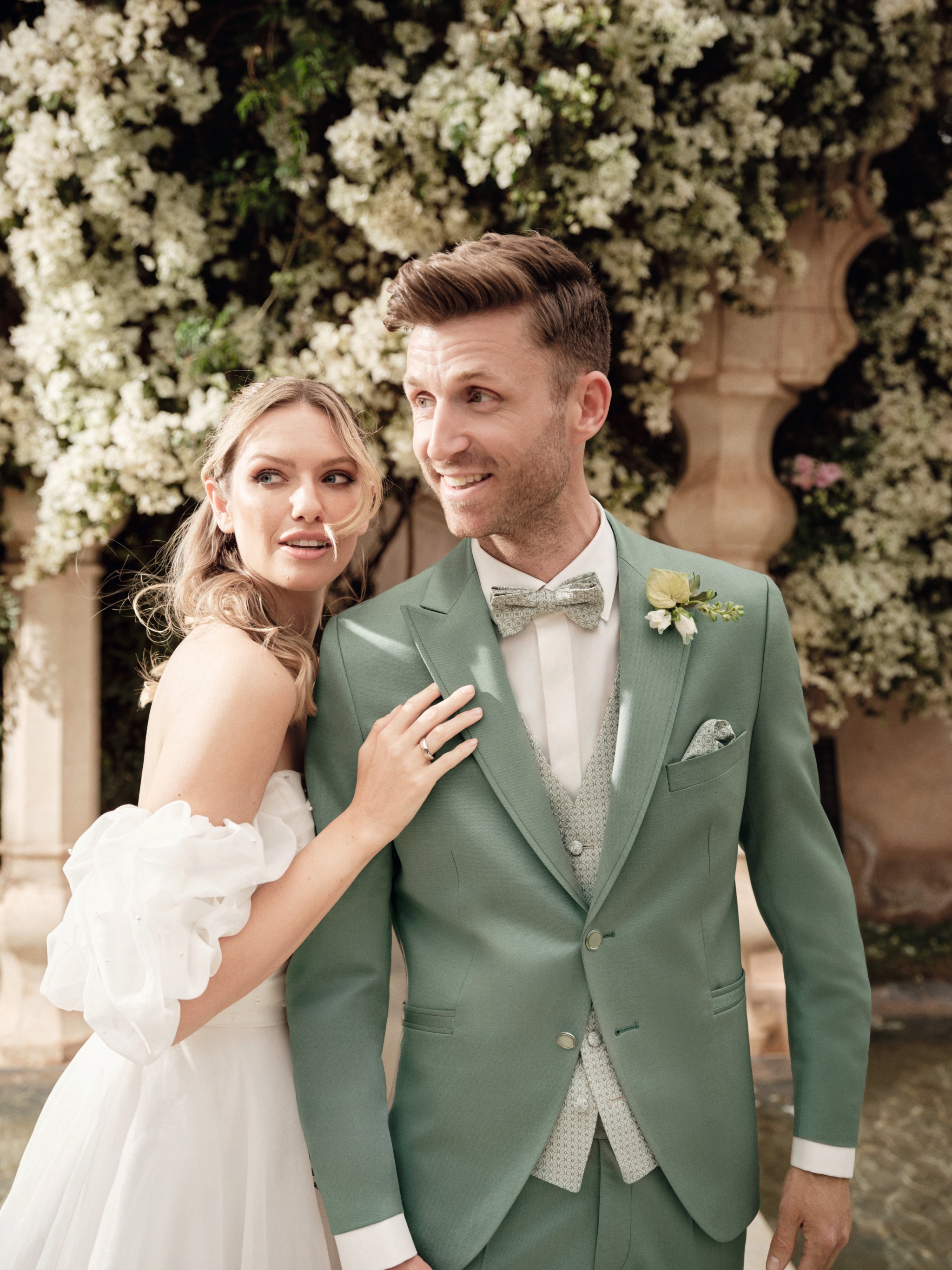 hochzeitsrausch Brautmoden - Brautkleid mit Off-Shoulder-Ärmeln und grüner Bräutigam-Anzug vor blühender Kulisse